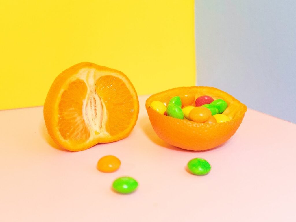 Orange cut in half. One side is filled with candy. On a tri-color muted color backdrop: yellow, pink, and blue.