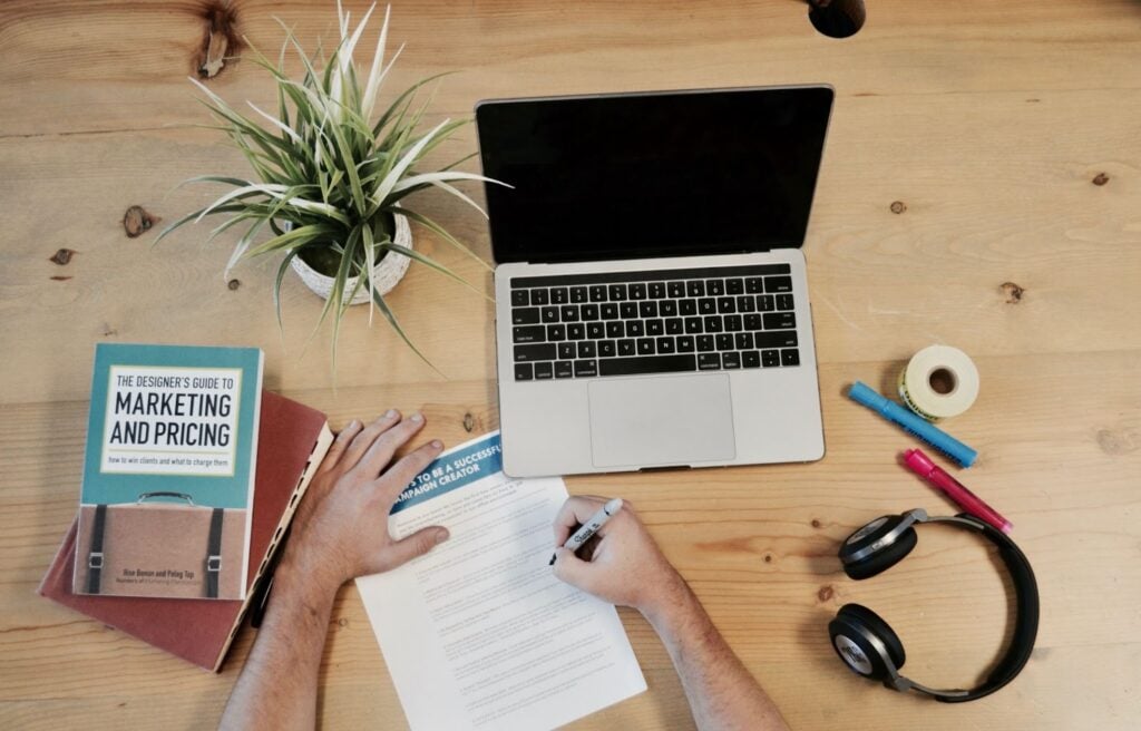 Person checking a a document on a desk with a laptop, marketing book, and headphones.